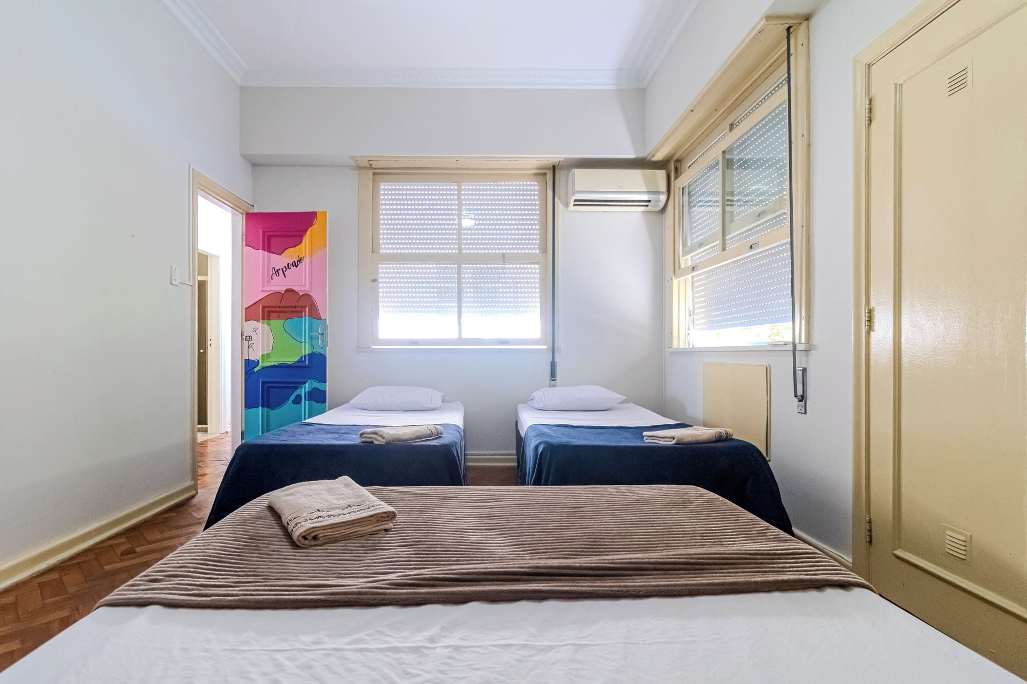 A room with two single beds, a large bed, a colorful artwork on the wall, and an air conditioning unit.