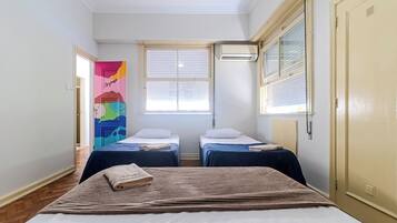 A room with two single beds, a large bed, a colorful artwork on the wall, and an air conditioning unit.