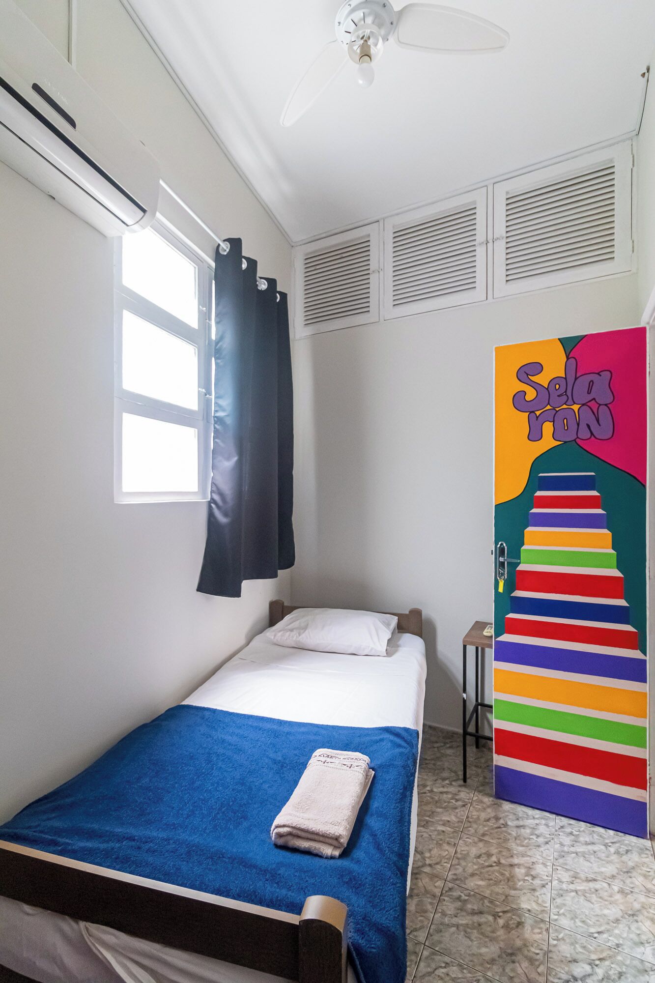 Quarto pequeno com cama de solteiro, colcha azul, almofadas brancas, cortina preta, ventilador de teto e um quadro colorido na parede.