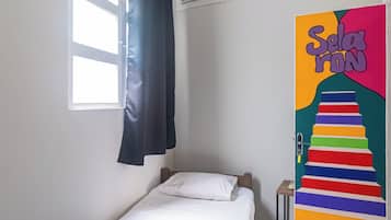 Quarto pequeno com cama de solteiro, colcha azul, almofadas brancas, cortina preta, ventilador de teto e um quadro colorido na parede.