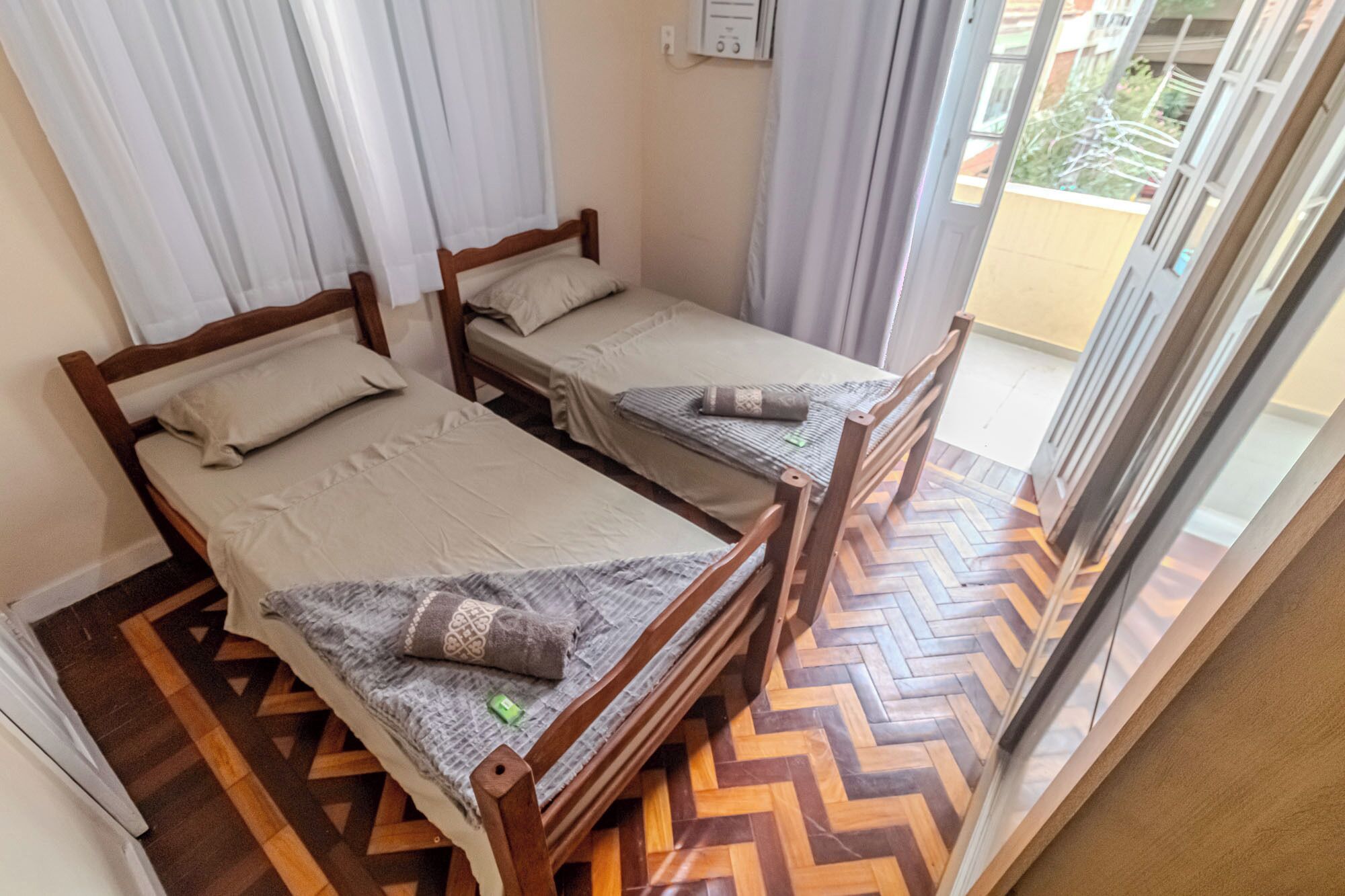 A room with two wooden beds, each with a pillow and a blanket. The room has a herringbone patterned floor and a sliding glass door leading to an outdoor area.