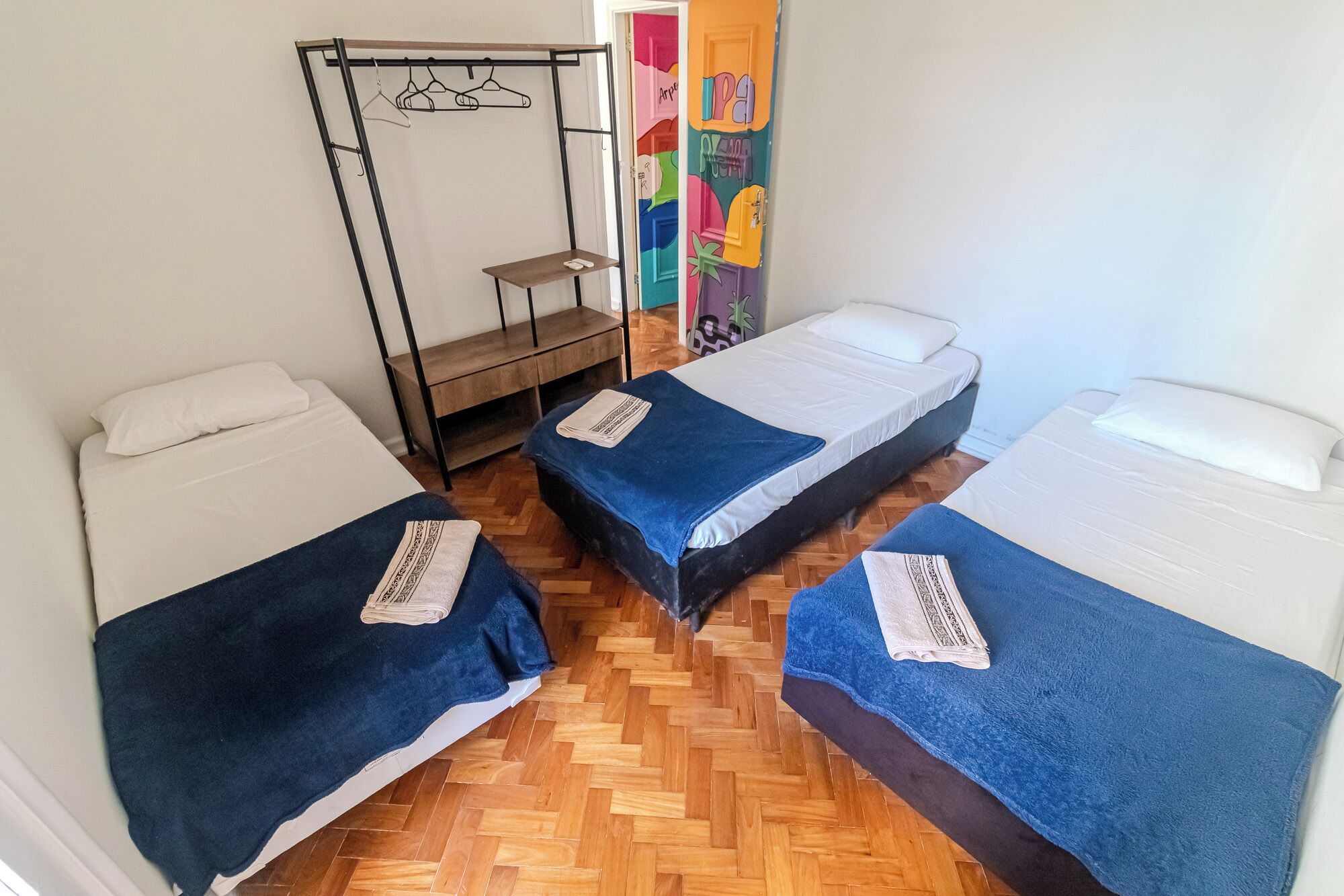 Une chambre avec trois lits simples, un plancher en bois, un portant métallique avec des cintres et une porte colorée.
