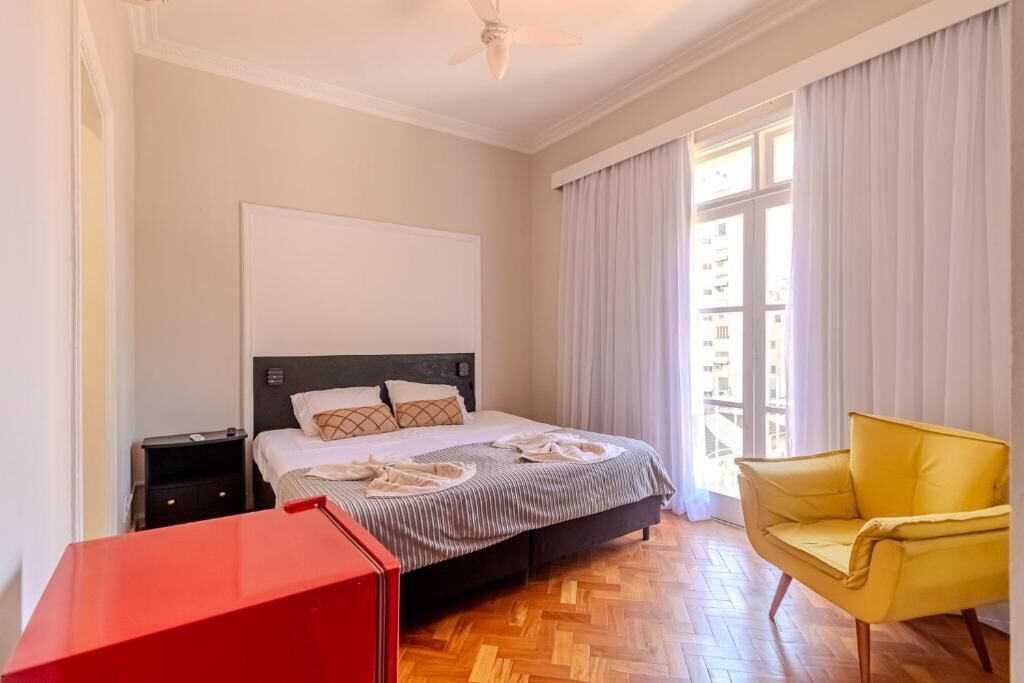 Une chambre d’hôtel avec un lit, une table de chevet, une table rouge, un fauteuil jaune et de grandes fenêtres avec des rideaux.