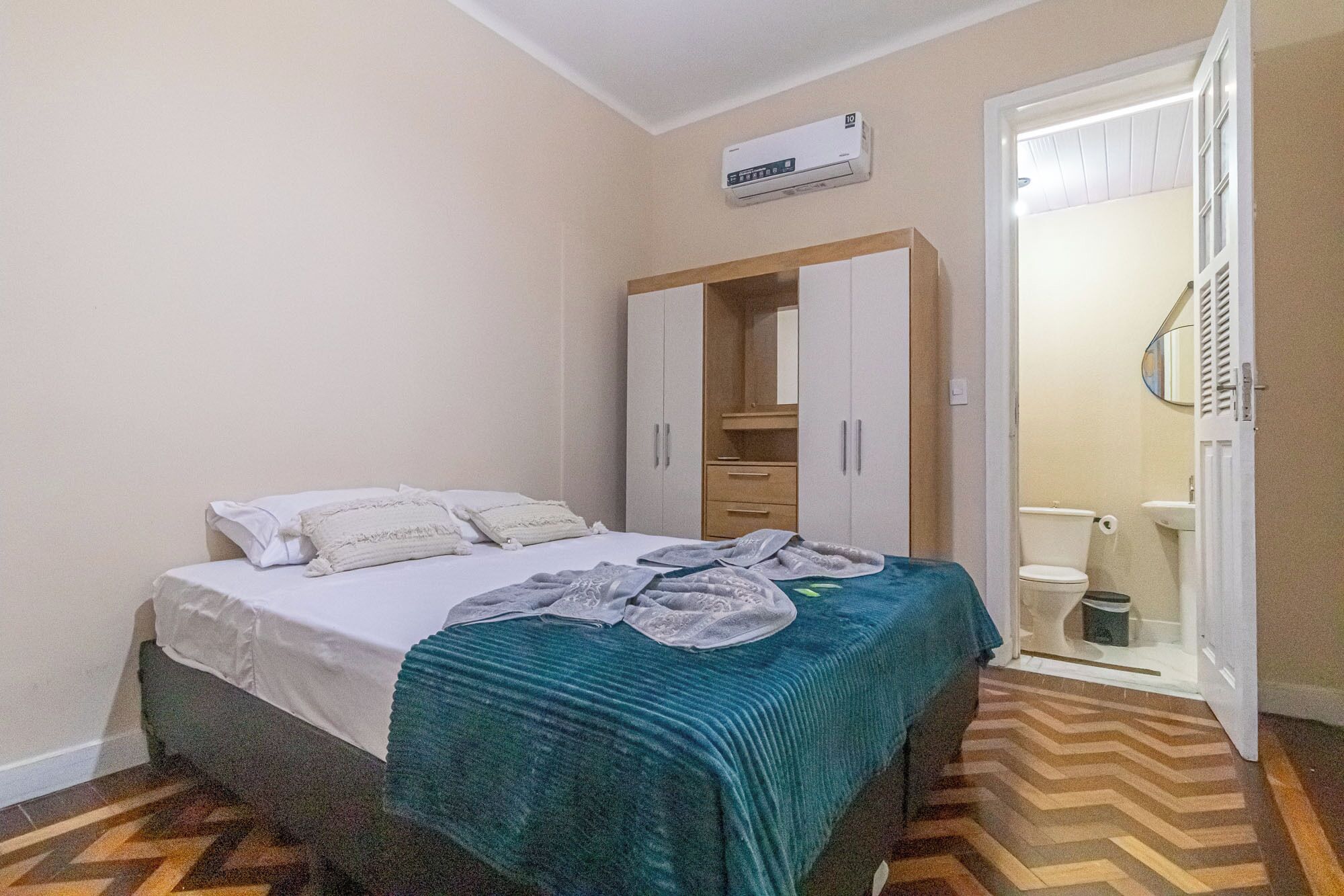 Une chambre à coucher avec un lit, une armoire, un miroir et une salle de bain visible à travers une porte ouverte.