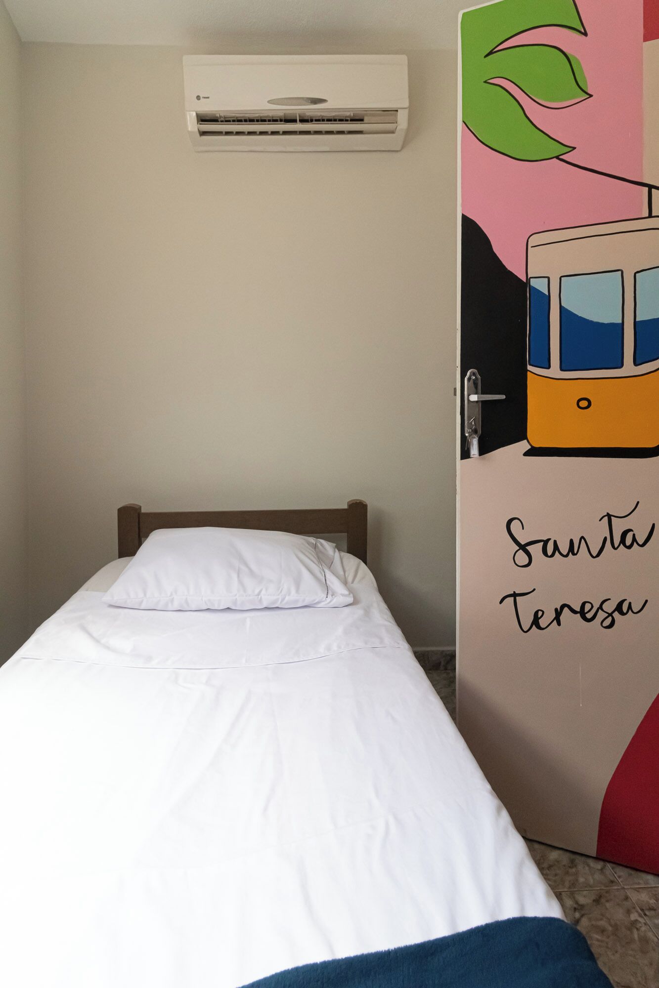 A single bed with white bedding, a wall-mounted air conditioner, and a door with a colorful mural.