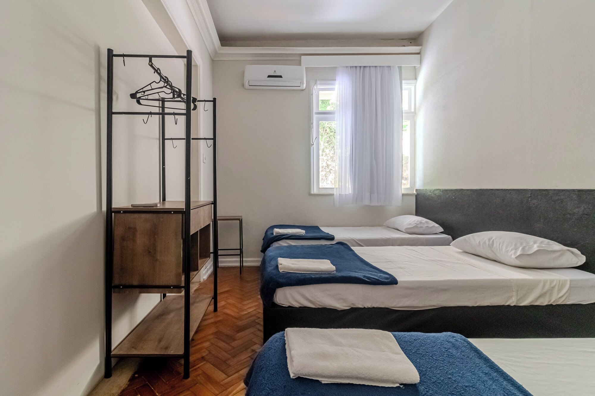 Une chambre de dortoir avec trois lits, un banc en bois et un étendoir en métal avec des cintres.