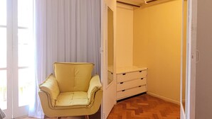 Une pièce avec un fauteuil jaune, un parquet en bois disposé en chevrons, un téléviseur fixé au mur et un dressing doté d’un miroir et de tiroirs.