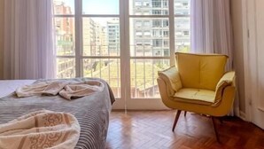 Une chambre à coucher avec un lit, un fauteuil jaune et de grandes fenêtres avec des rideaux.