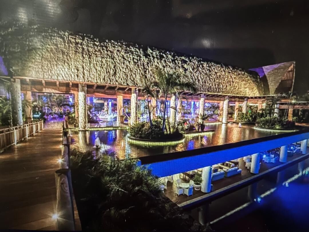 Un grand bâtiment illuminé, avec un bassin décoratif devant, entouré de palmiers et agrémenté d’espaces de détente en plein air.