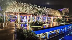 Un grand bâtiment illuminé, avec un bassin décoratif devant, entouré de palmiers et agrémenté d’espaces de détente en plein air.
