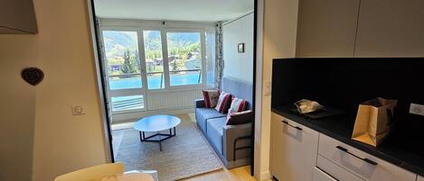 A compact kitchen with a dining area, a sofa with cushions, and a view of a lake and mountains through large windows.
