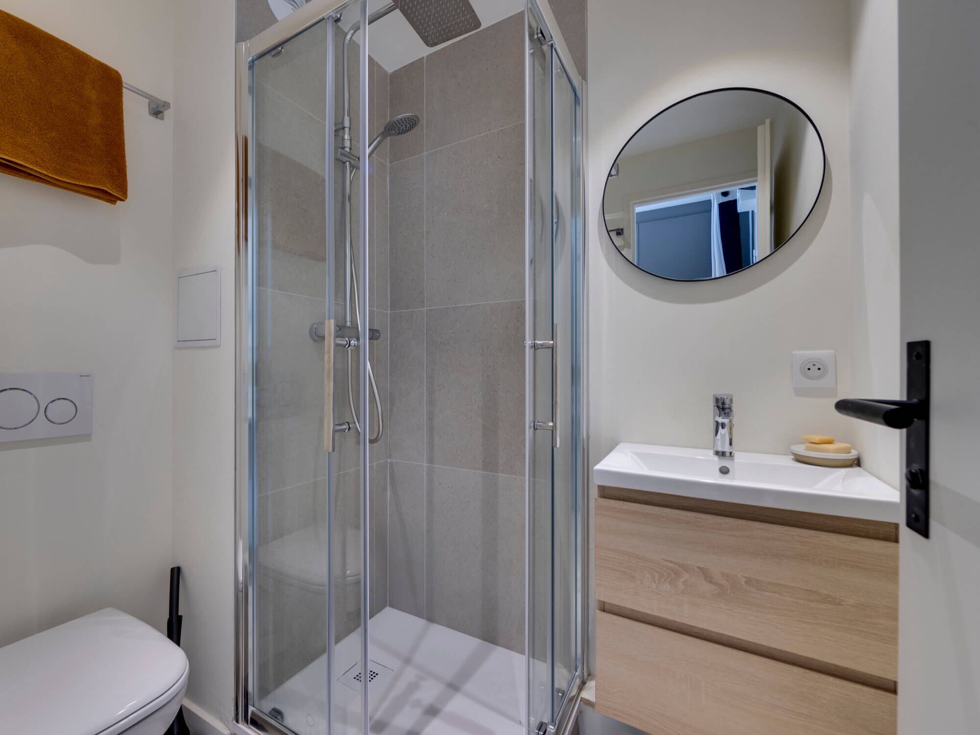 A modern bathroom with a glass-enclosed shower, a white sink with a wooden vanity, and a round mirror on the wall.