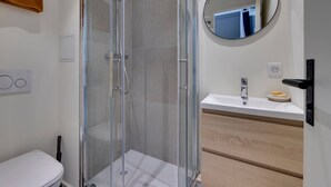Une salle de bain moderne avec une douche à l’italienne en verre, un lavabo blanc avec un meuble-vasque en bois et un miroir rond au mur.