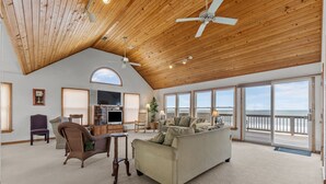 A spacious living room with a high ceiling, wooden beams, a large sofa, a television, and a view of the ocean through large windows. - 5854 - Erkkila. Relax at Erkkila, your perfect Outer Banks getaway near the beach. (Nags Head)