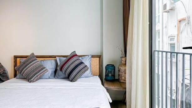 Un lit avec des coussins à motifs, une tête de lit en bois, un vase et une vue sur des immeubles par la fenêtre.