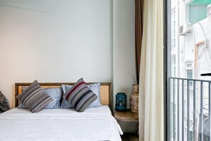 Sängyssä on kuviollisia tyynyjä, puinen pääty ja maljakko, ja ikkunasta avautuu näkymä rakennuksiin.