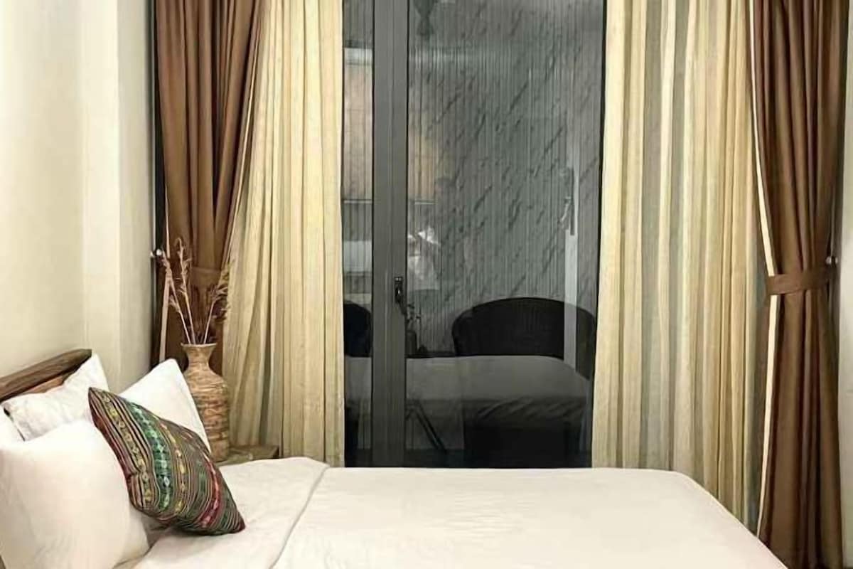 A hotel room with a bed, a vase with dried plants, and a view of a balcony with a chair.