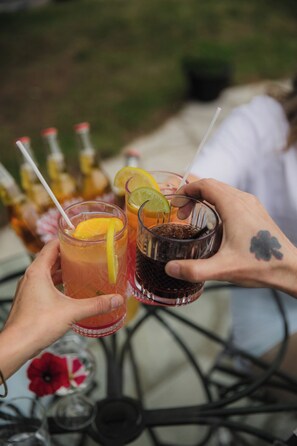 Deux mains tenant des verres remplis de boissons, l’un muni d’une paille et l’autre d’une fourchette, sur une table avec des bouteilles en arrière-plan.