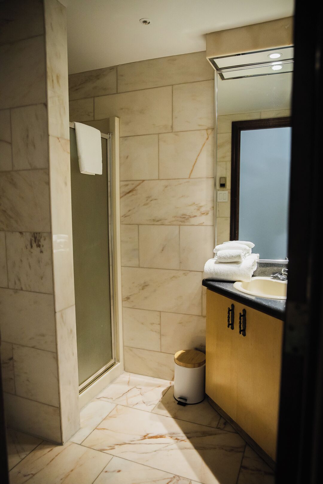 Baño con cabina de ducha de vidrio, lavamanos con espejo y un cubo de basura.