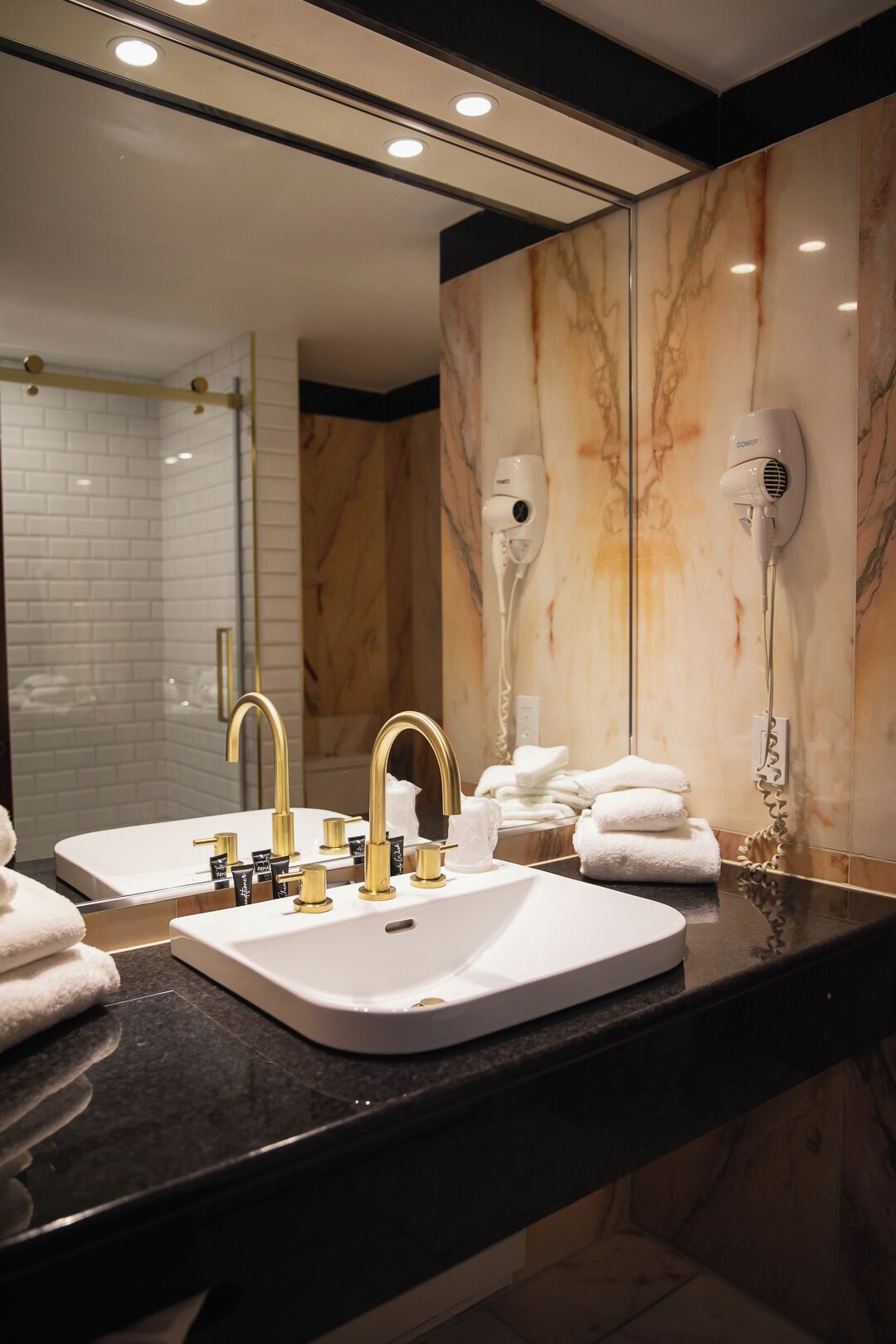 A bathroom with a large mirror, two sinks with gold faucets, a marble wall, and two hair dryers mounted on the wall.