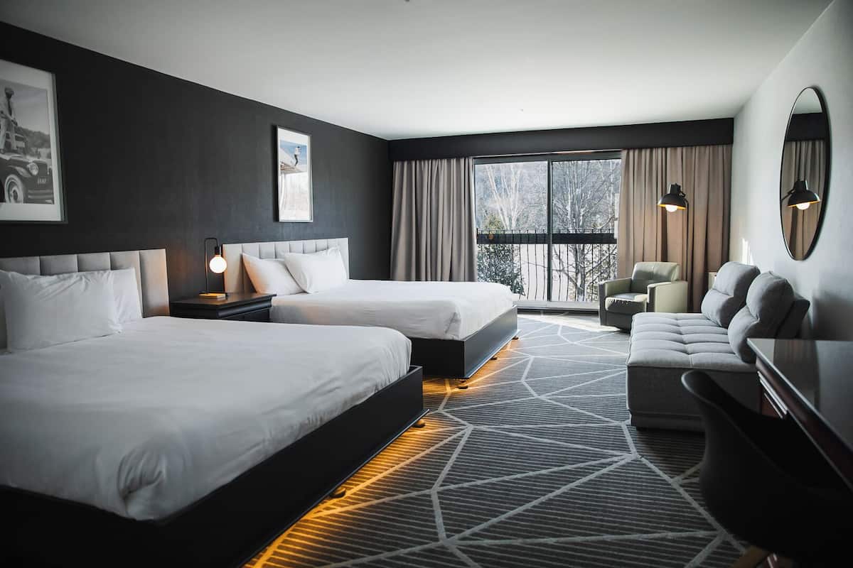 A modern hotel room with two beds, a sofa, a desk, and a large window with curtains.