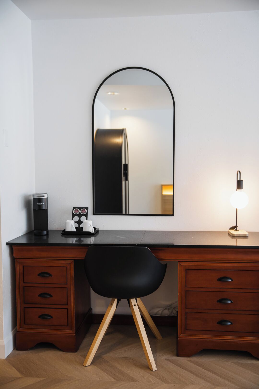 Una habitación de hotel moderna con un escritorio de madera, una silla negra, un espejo, una cafetera y una lámpara.