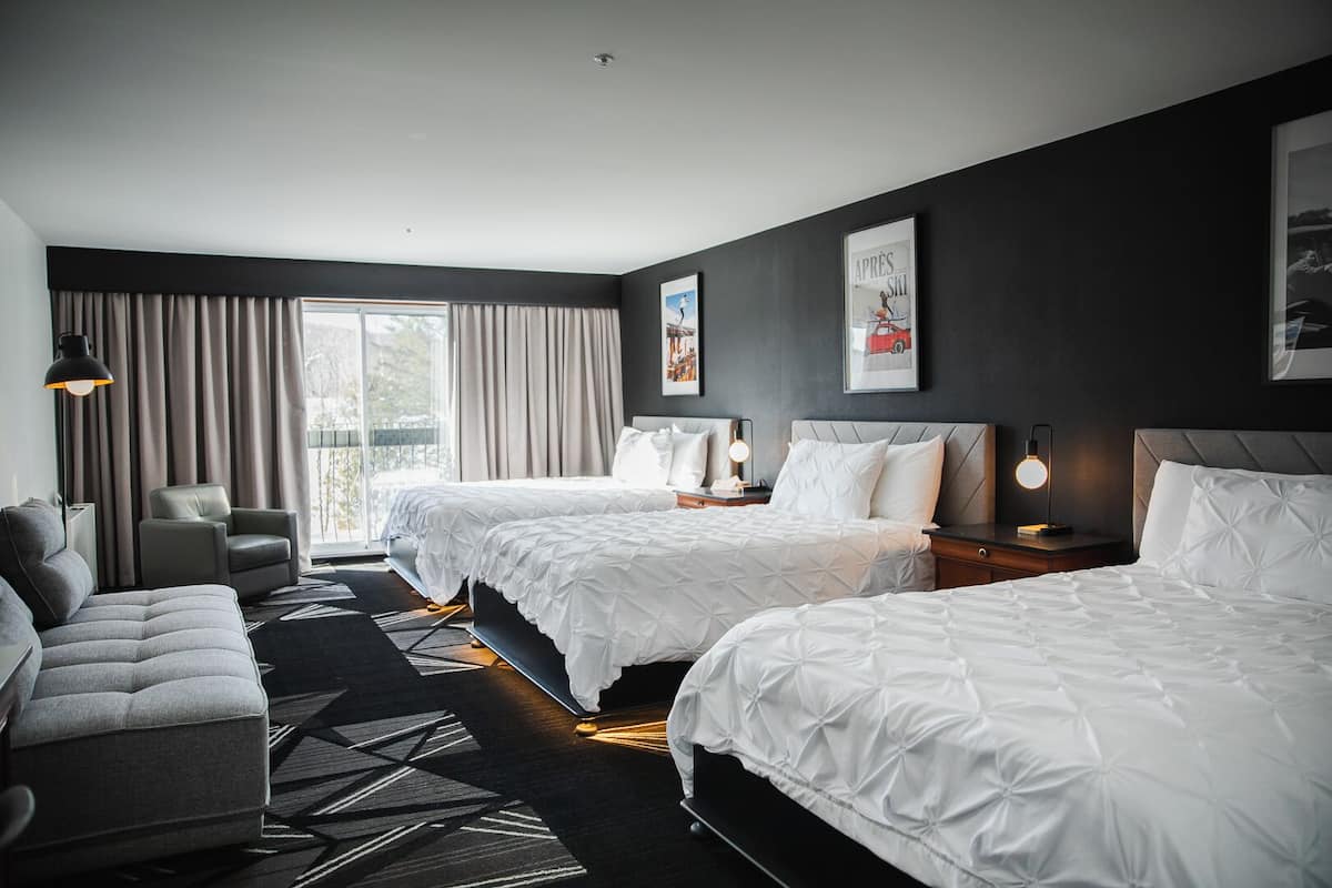 A modern hotel room with two beds, a sofa, a desk, and a large window with curtains.
