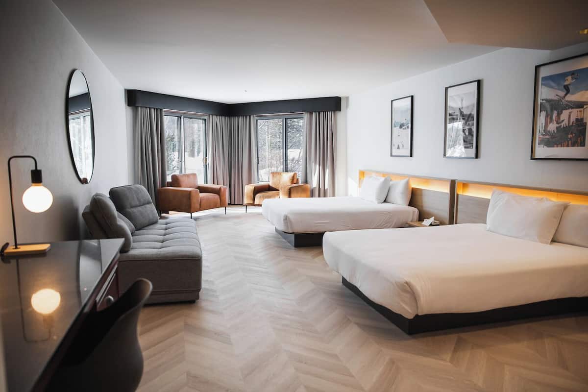 A modern hotel room with a sofa, two beds, a desk, and a large window with curtains.