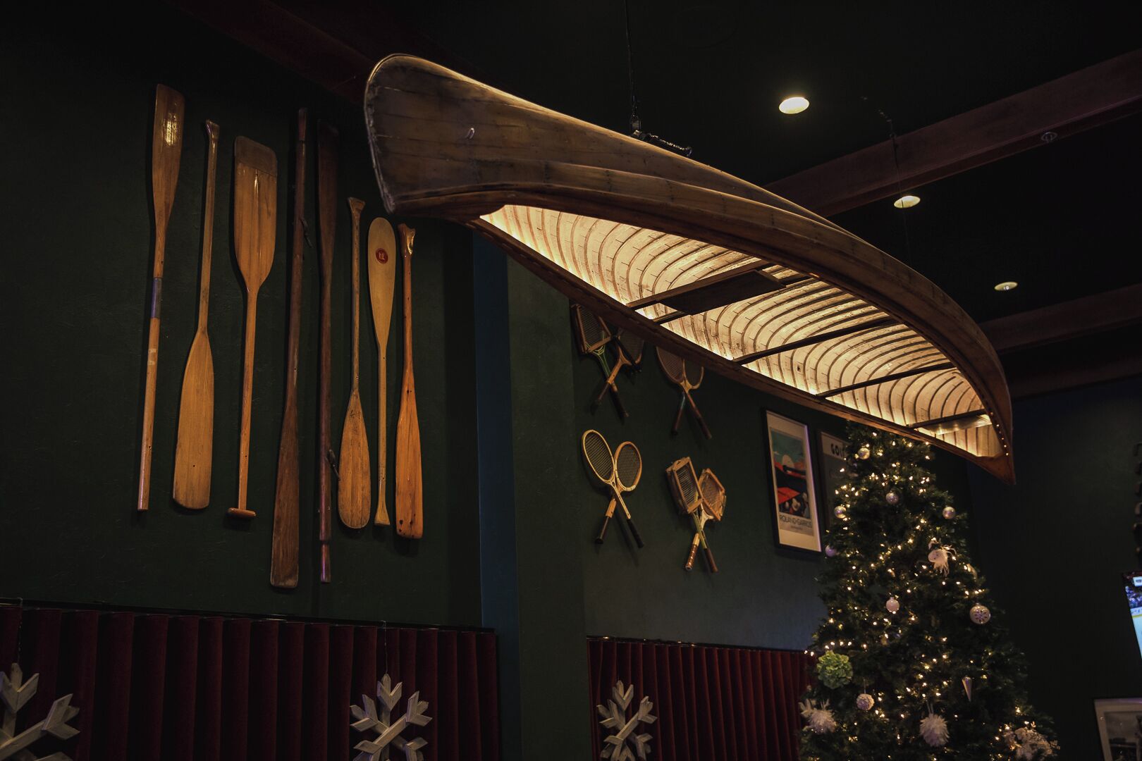 A room with a wooden canoe, various paddles, and tennis rackets hanging on the wall, and a Christmas tree.
