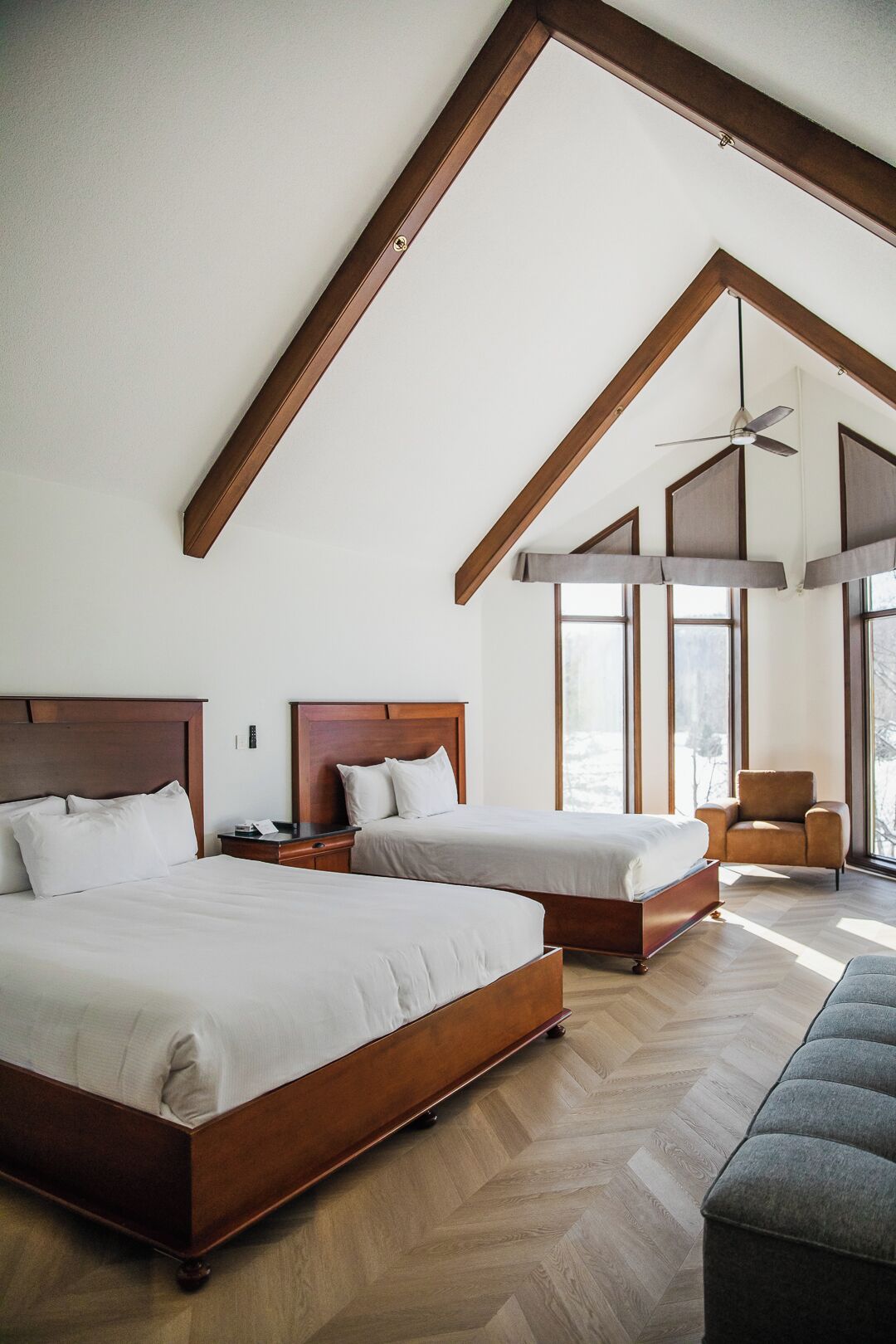 A spacious bedroom with two large beds, wooden headboards, a ceiling fan, and large windows allowing natural light.