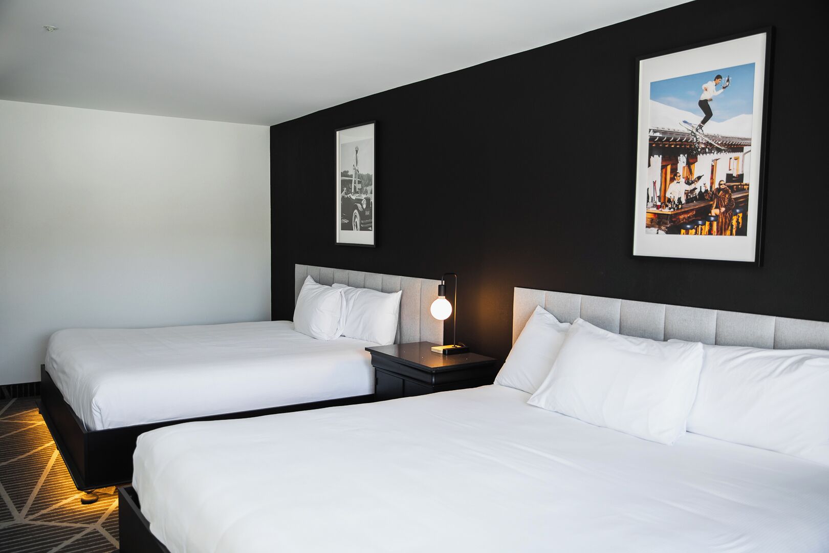 Habitación de hotel con dos camas individuales, una mesita de noche con lámpara y dos cuadros en la pared.