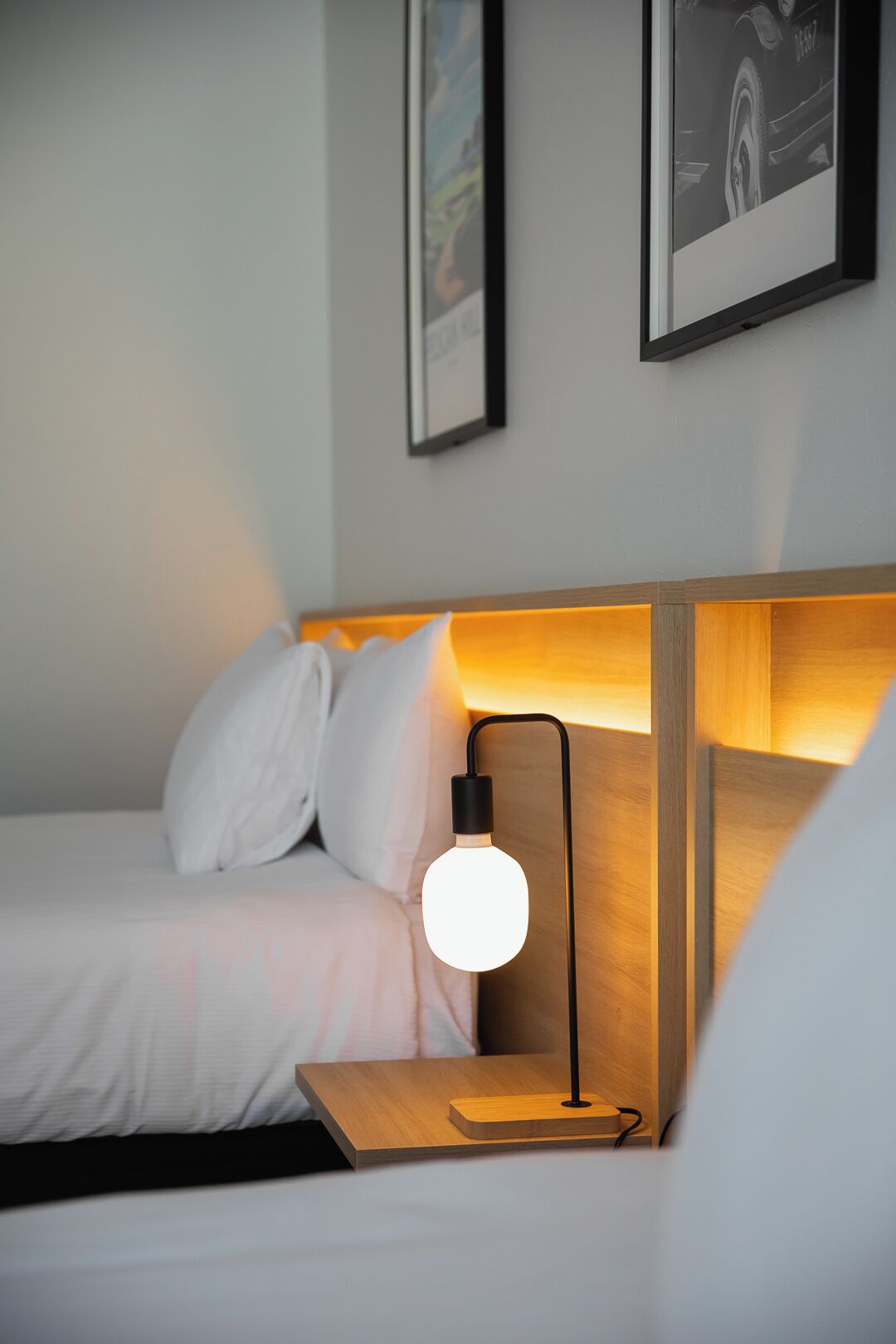 A neatly made bed with white linens and pillows, a bedside table with a lamp, and framed pictures on the wall.