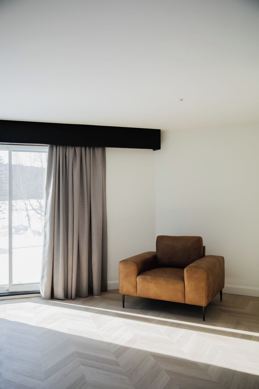 A modern living room with a large window, a brown sofa, and a herringbone-patterned floor.