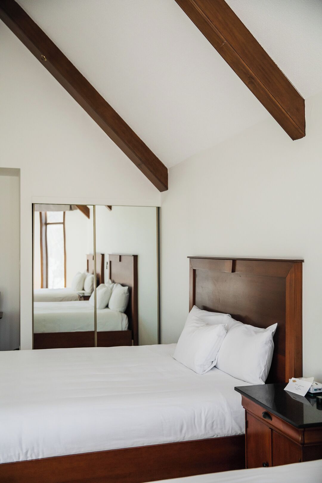 Een slaapkamer met een groot bed, een houten hoofdbord, een nachtkastje en een spiegel die de kamer weerkaatst.