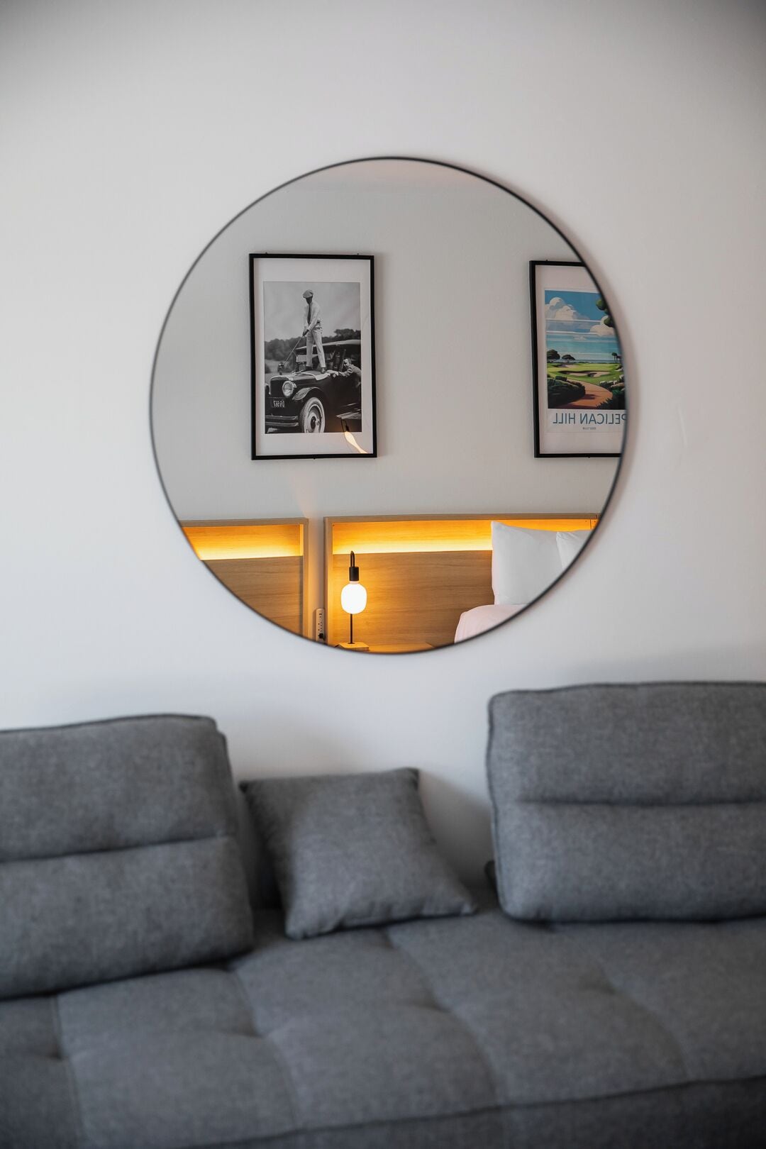 Una sala de estar moderna con un sofá gris, un espejo redondo que refleja una cama y una lámpara, y dos cuadros enmarcados en la pared.