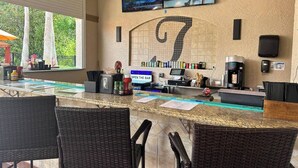 A bar area with wicker chairs, a counter with a coffee machine, and a TV screen displaying a sports event. - Hideaway Gem near Disney & golf. Top floor bonus (Davenport)