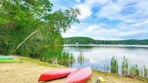 A lakeside setting with a canoe and paddle on the sand, surrounded by trees and a calm body of water under a partly cloudy sky. - Stunning Sterling Sanctuary: Unwind on the Water! (Sterling)