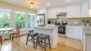A modern kitchen with white cabinets, a central island with stools, a dining area with a table and chairs, and large windows offering a view of greenery. - Stunning Sterling Sanctuary: Unwind on the Water! (Sterling)
