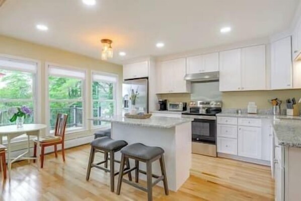 A modern kitchen with white cabinets, a central island with stools, a dining area with a table and chairs, and large windows offering a view of greenery. - Stunning Sterling Sanctuary: Unwind on the Water! (Sterling)