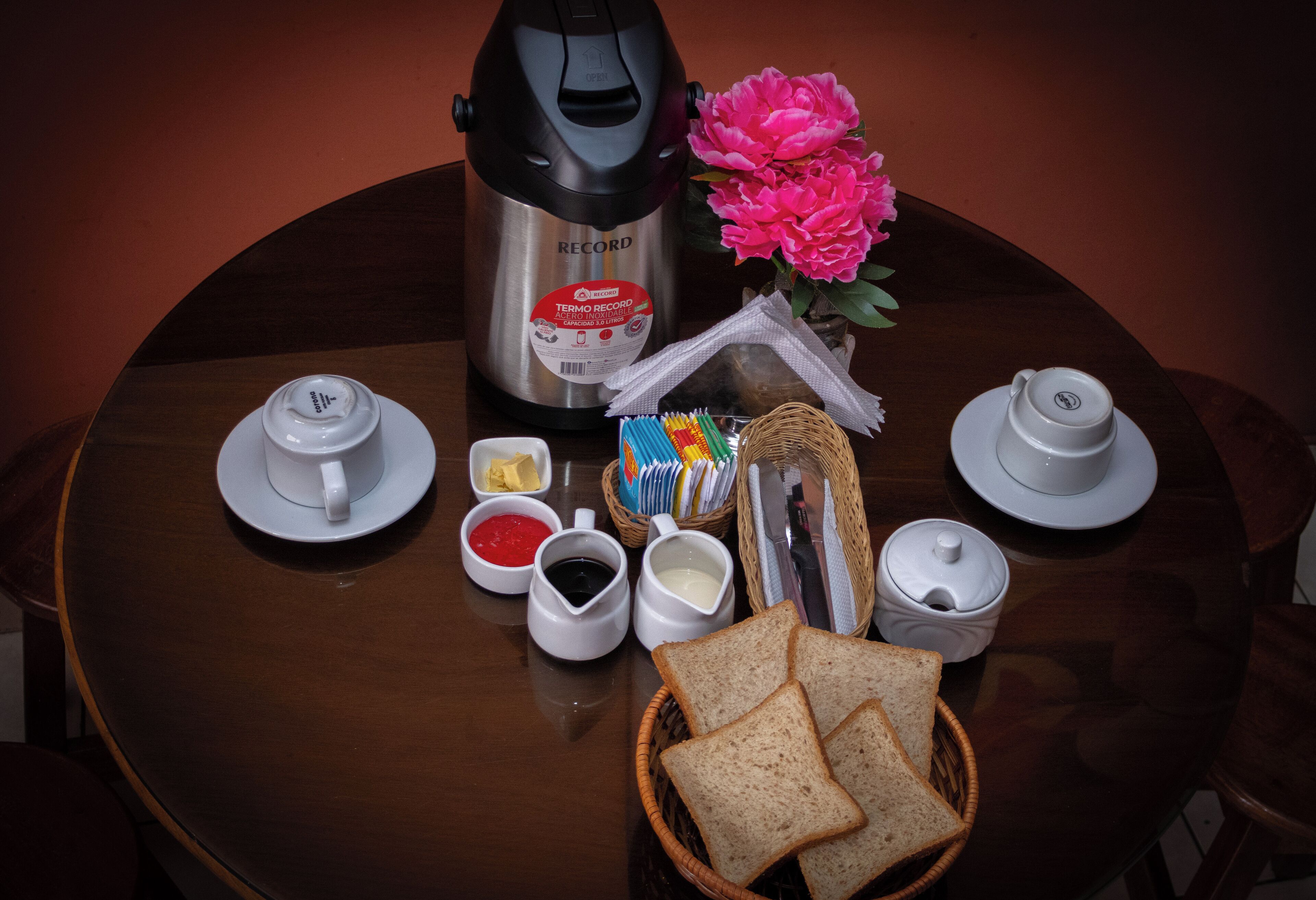 Une table en bois dressée pour le petit-déjeuner, avec une machine à café, des tasses à thé, du pain, des condiments et une fleur décorative.
