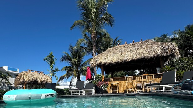 Une piscine aux eaux cristallines, bordée de chaises longues et abritée par une structure au toit de chaume, le tout entouré de palmiers.