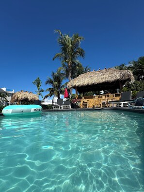 Une piscine aux eaux cristallines, bordée de chaises longues et abritée par une structure au toit de chaume, le tout entouré de palmiers.