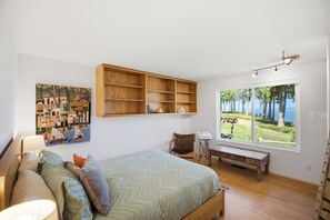 Un dormitorio con cama, estante de madera, ventana con vistas y un cuadro en la pared.
