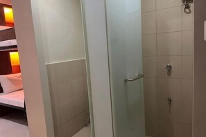 Une pièce dotée d’une porte en verre donnant sur une zone de douche, d’un lit avec du linge de lit blanc et d’une lampe fixée au mur.
