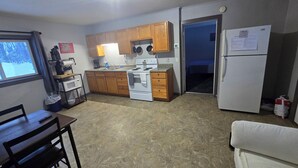 A compact kitchen with wooden cabinets, a white stove, and a refrigerator. There is a dining table with chairs and a window with a view of trees. - WP3 MTU & Keweenaw Base Full Kitchen + WiFi (Calumet)