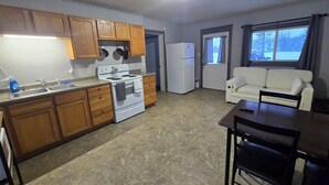 A compact kitchen with wooden cabinets, a white stove, and a refrigerator. There is a dining area with a table and chairs, and a white sofa. - WP3 MTU & Keweenaw Base Full Kitchen + WiFi (Calumet)
