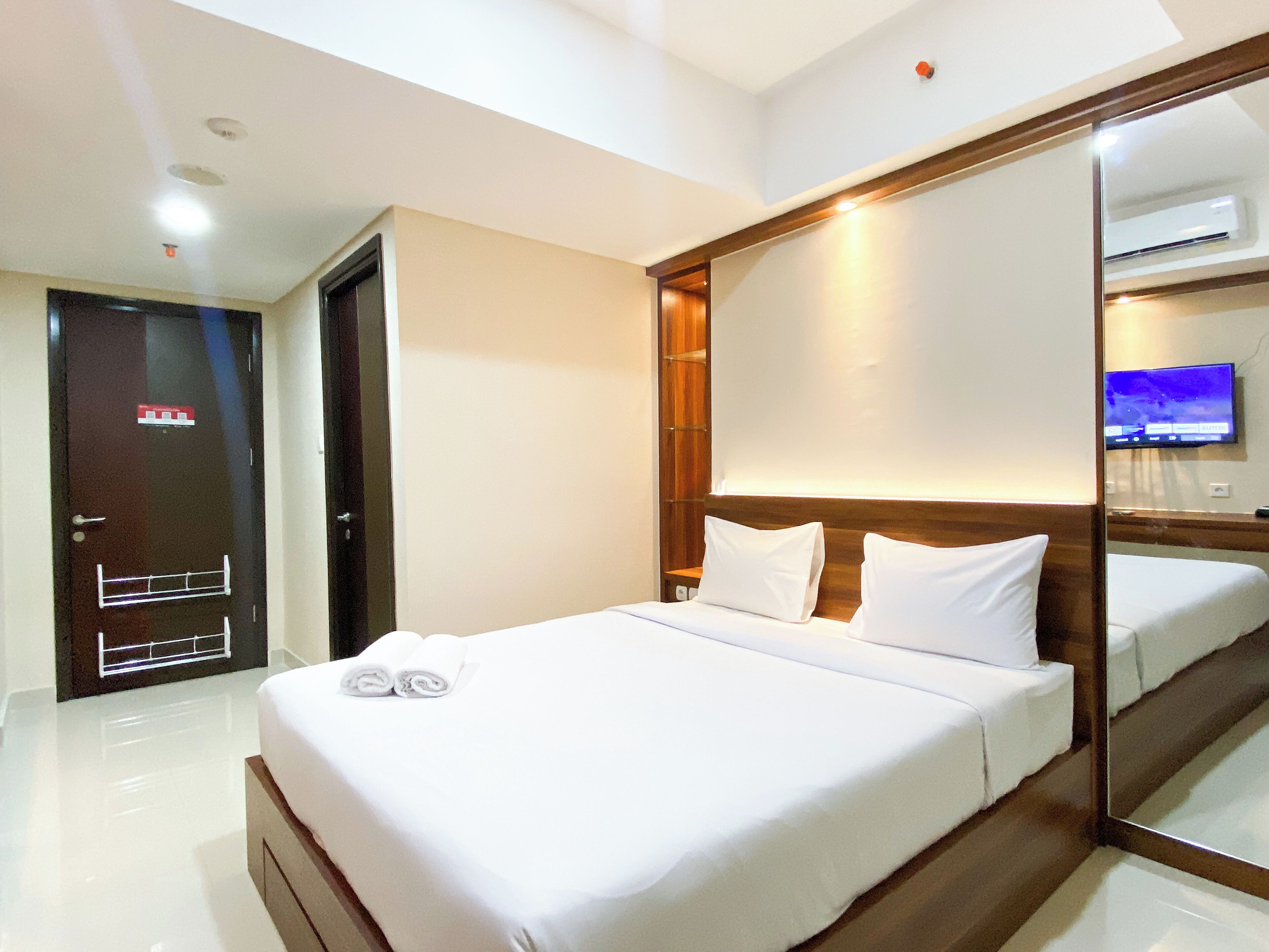 Habitación de hotel con una cama grande, cabecera de madera, espejo, televisor y puerta con cámara de seguridad.