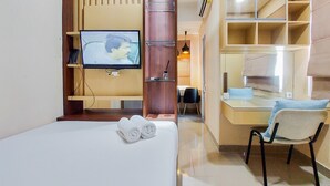 A hotel room with a bed, a TV mounted on a wooden panel, a desk with a chair, and a mirror. - Strategic And Comfy Studio Apartment B Residence Near Bsd (Pagedangan)