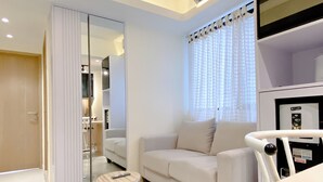 A modern living room with a sofa, a coffee table, and a small side table. There is a large mirror on one wall and a window with curtains. - Homey And Good Choice 2Br At Meikarta Apartment (Cikarang)