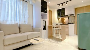 A modern living room with a sofa, a small round table, and a kitchen area with a fridge, a sink, and a stove. - Homey And Good Choice 2Br At Meikarta Apartment (Cikarang)